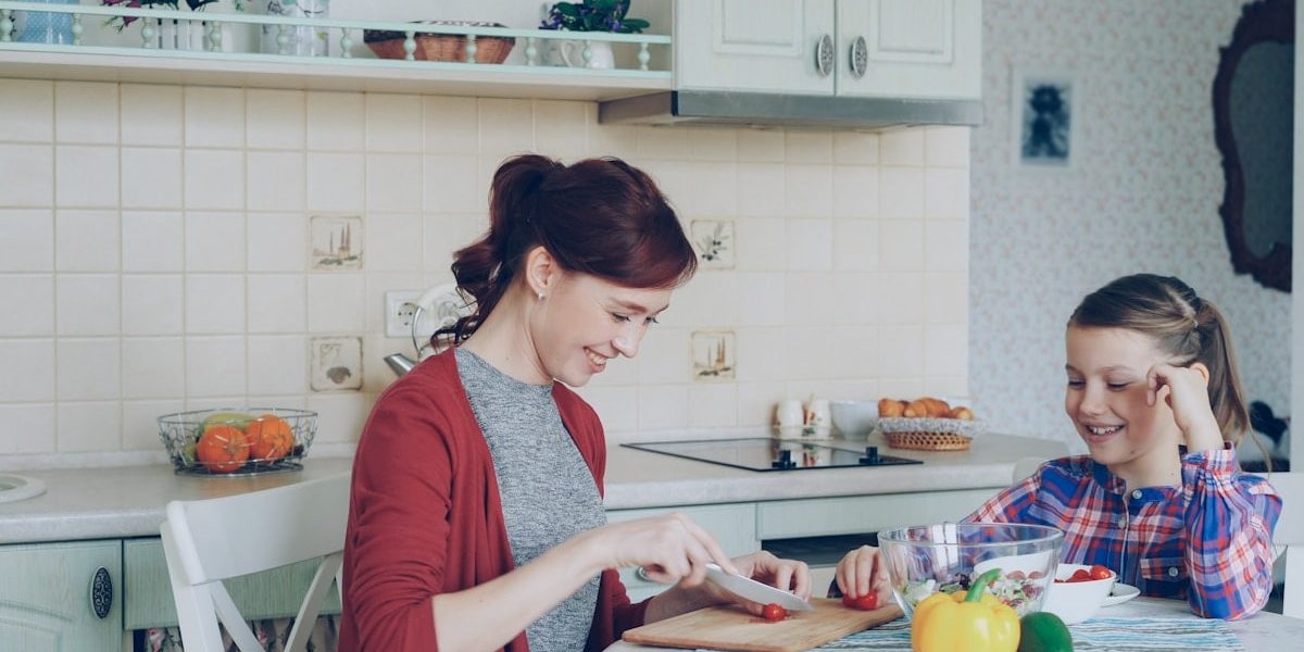 Happy young mother cooking vegetables cutting tomatoes and talking to her cheerful daughter in the kitchen at home. Family, cook, and people concept