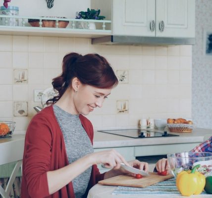 Happy young mother cooking vegetables cutting tomatoes and talking to her cheerful daughter in the kitchen at home. Family, cook, and people concept